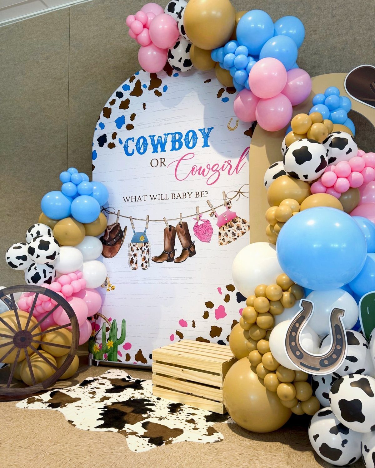 Western gender reveal backdrop with cowboy boots, cow-print balloons, and a wooden wagon wheel.