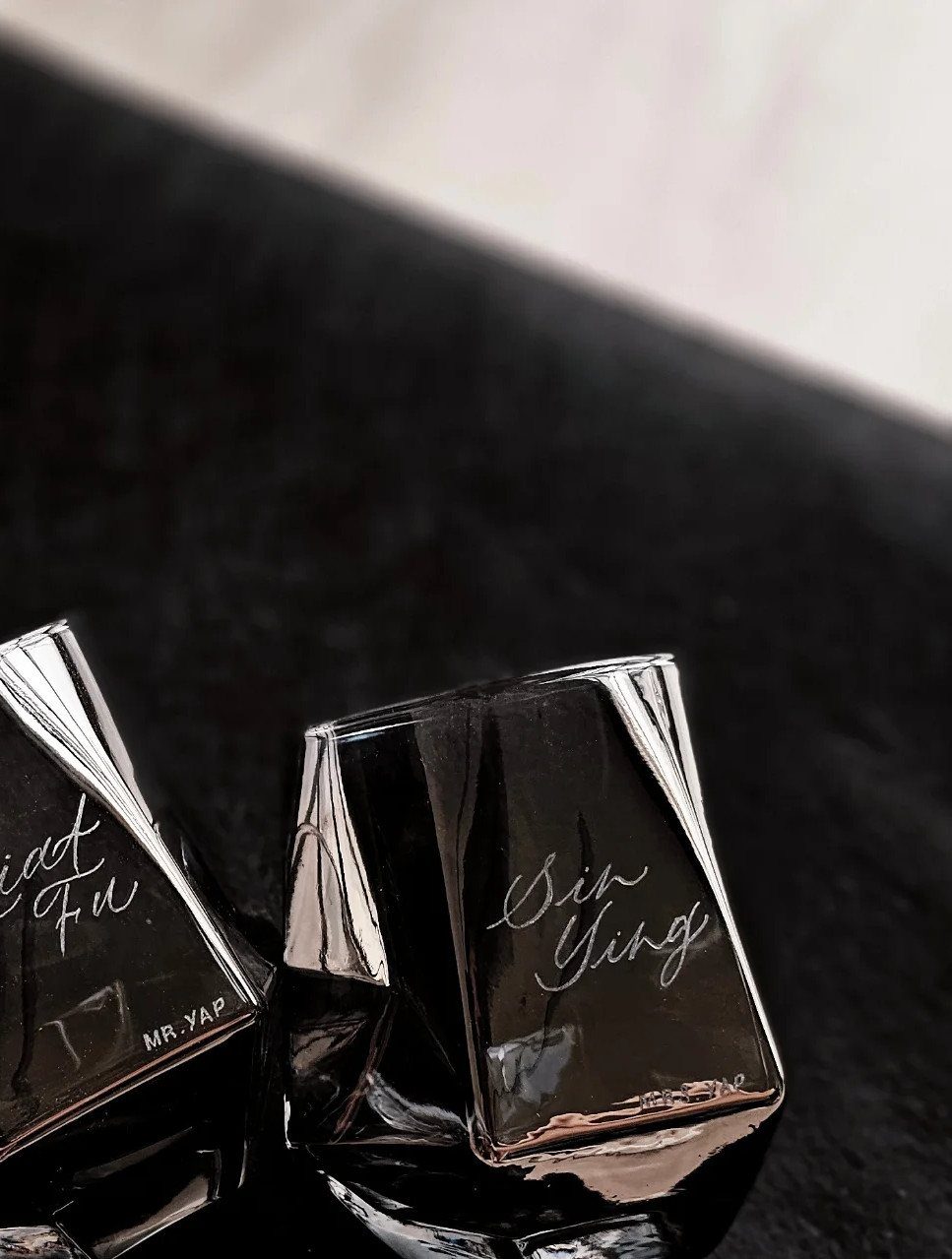Two black engraved square glasses with cursive names on a dark surface.