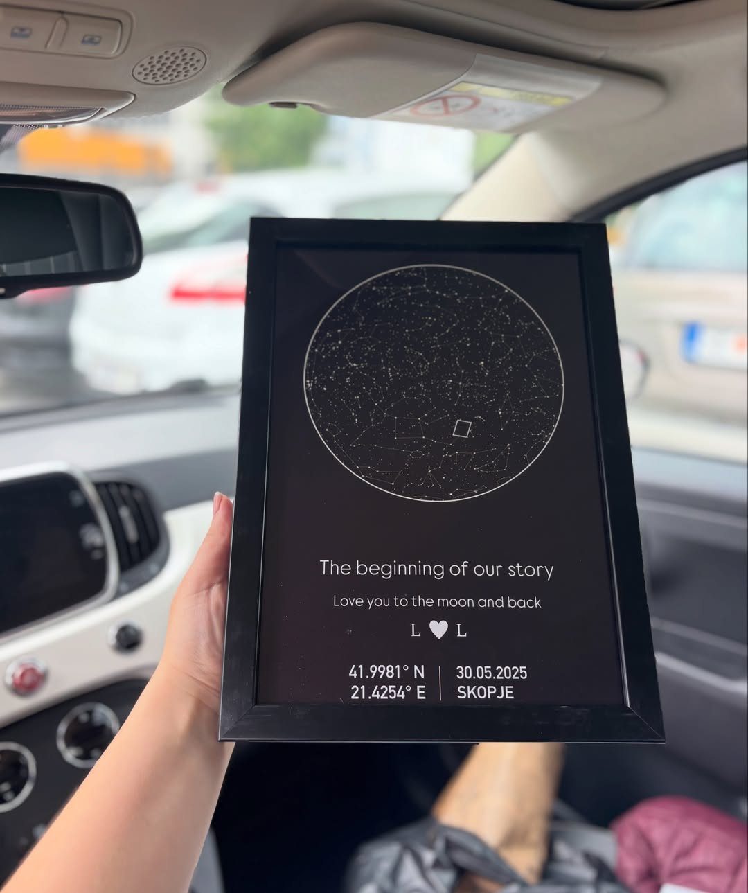 Black framed personalized star map held inside a car with white text below.