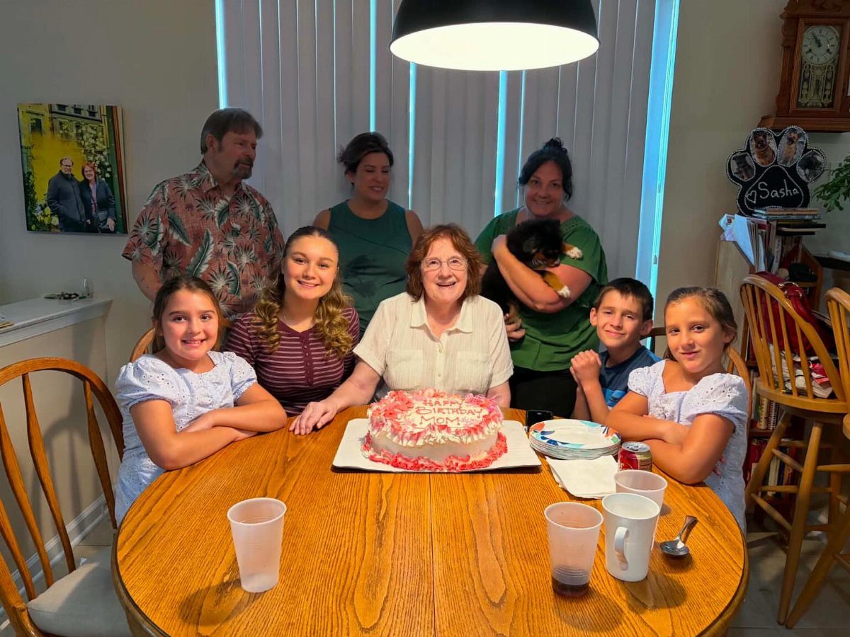 Family gathering around a wooden table with a birthday cake for an older woman.