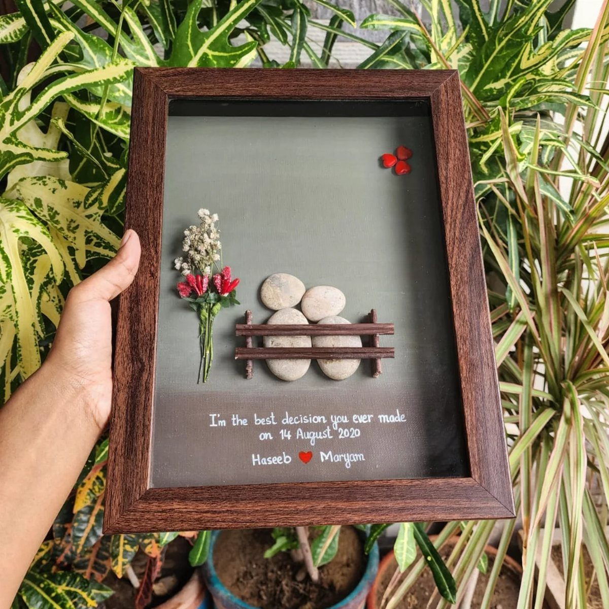 Hand holding a framed 3D shadow box with pebbles, flowers, and romantic text.