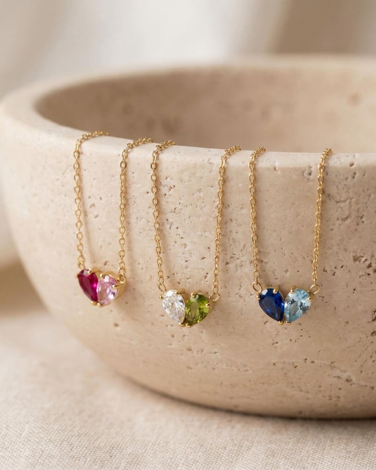 Three gold necklaces with colorful heart-shaped birthstone pendants draped over a stone bowl.