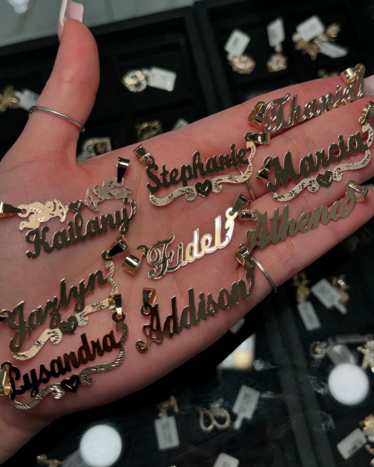Gold personalized name pendants held in a hand, featuring various cursive names.
