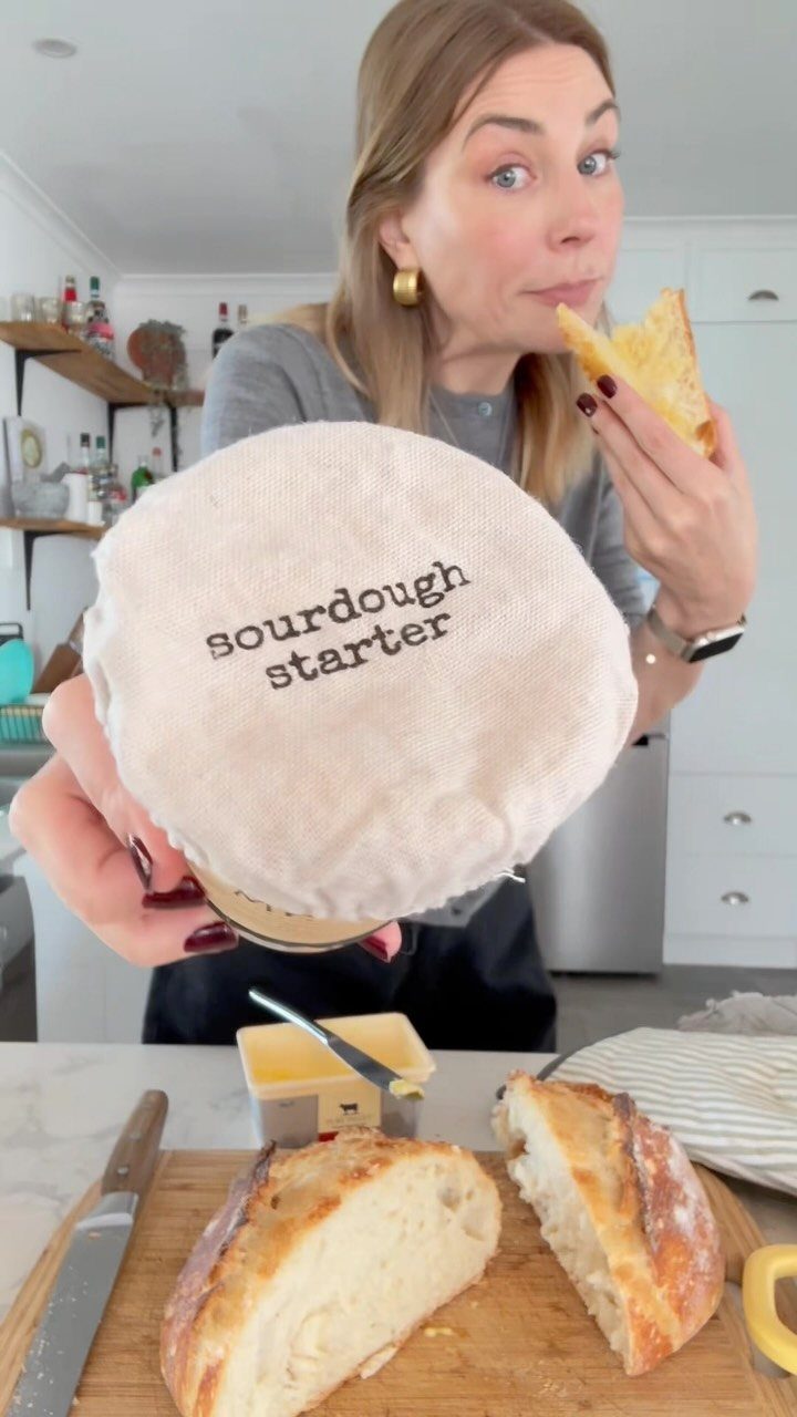 Woman holding a sourdough starter jar with fresh baked bread on a board.