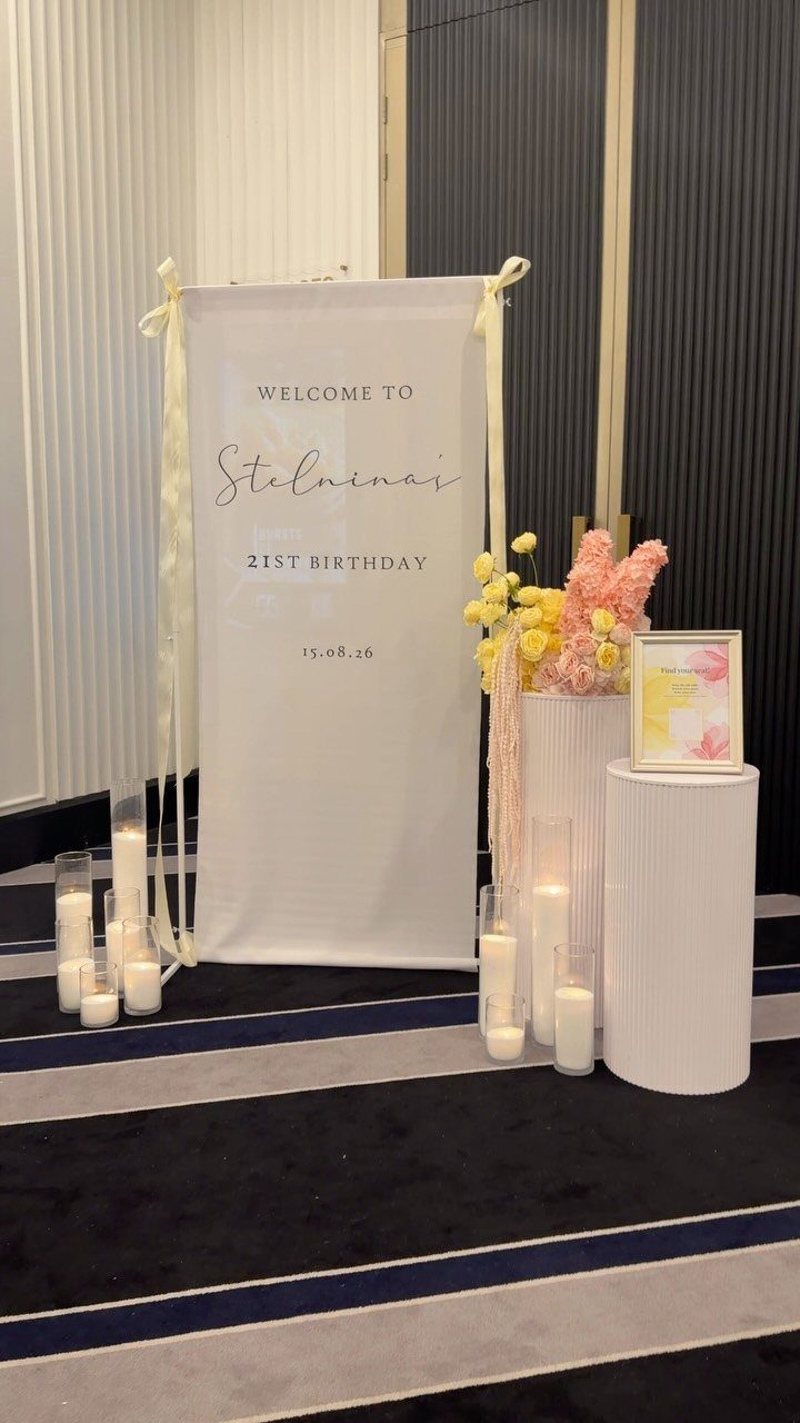 White 21st birthday welcome sign with gold text, flowers, and white pillar candles.