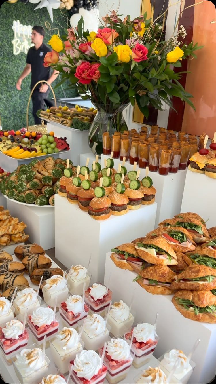 Catering spread with mini sliders, sandwiches, desserts, and a colorful floral bouquet.