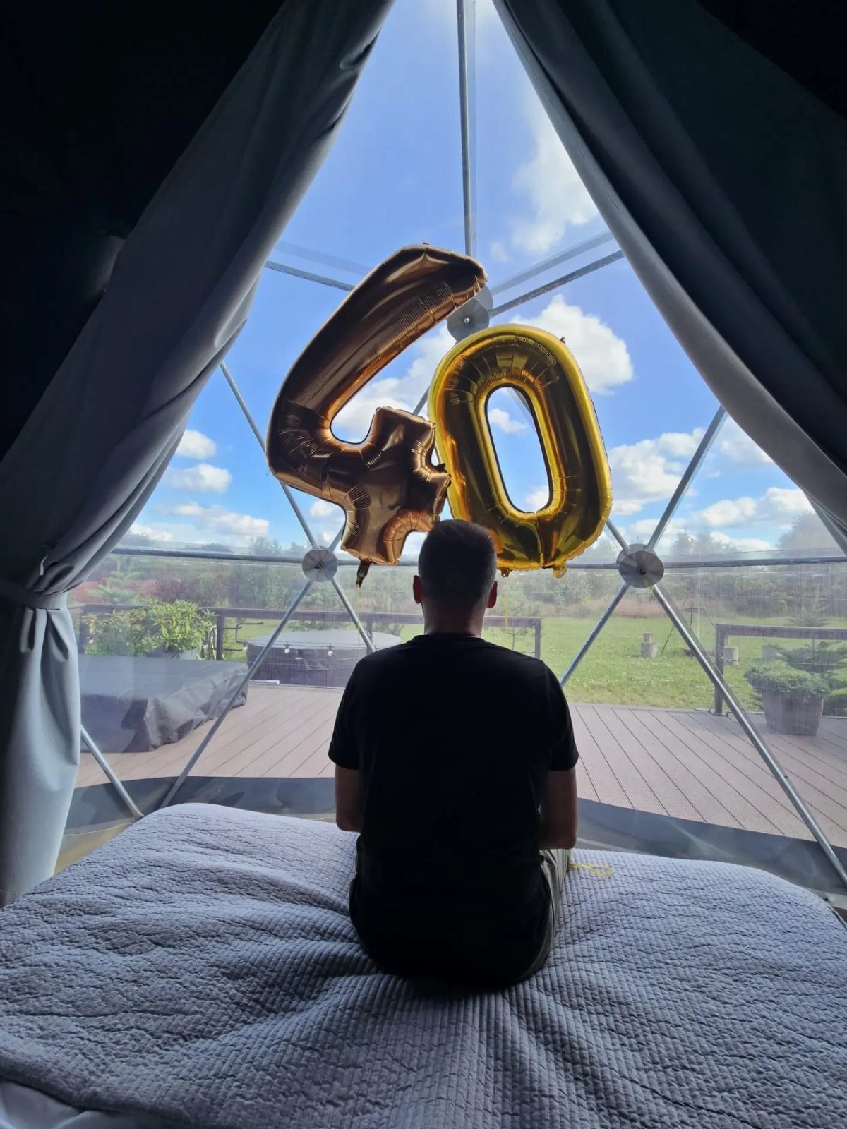 Man sitting in a transparent dome tent with gold 40th birthday balloons.