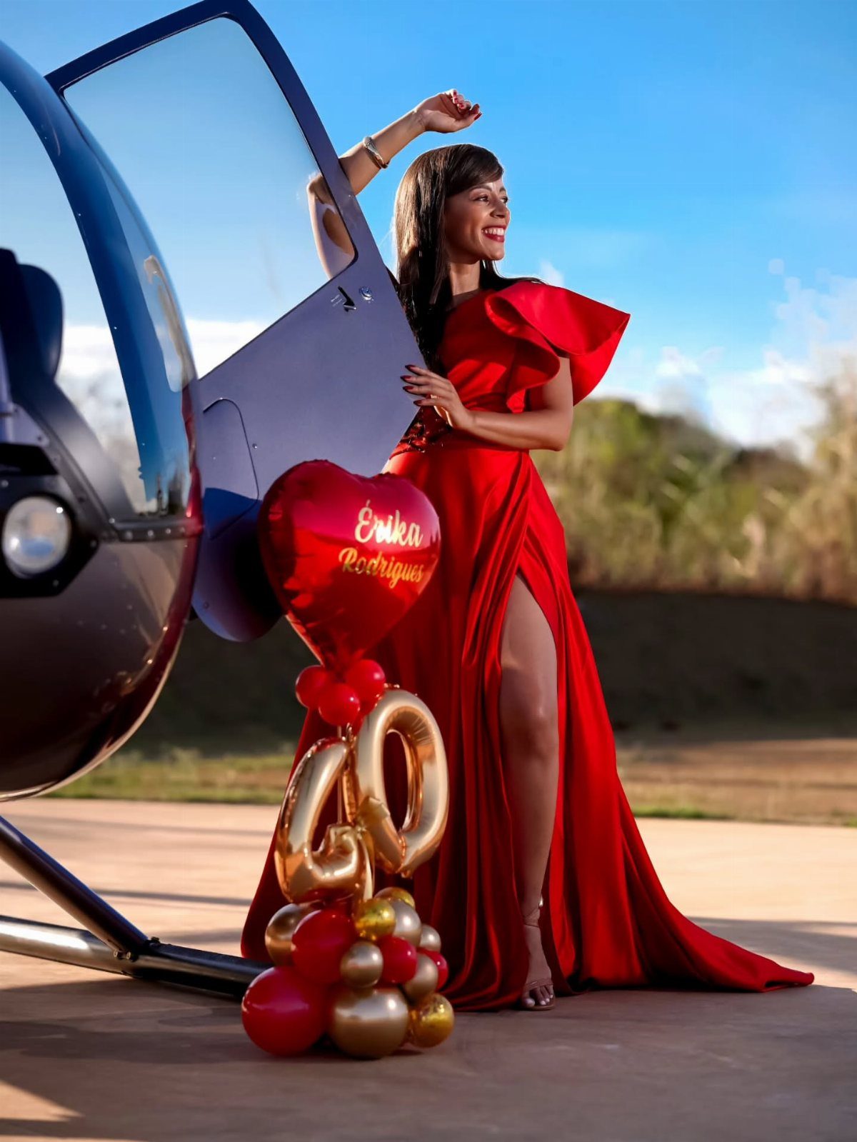 Woman in red gown posing with 40th birthday balloons next to a helicopter.