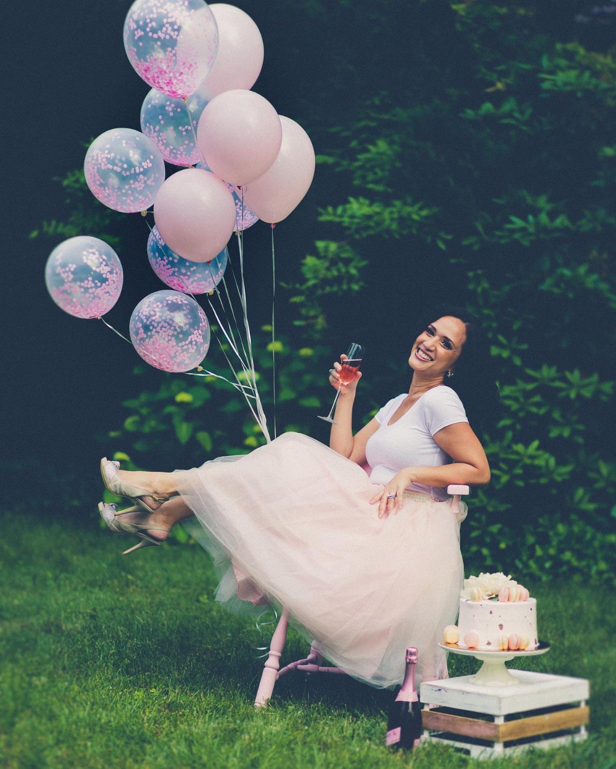 Woman in a pink tulle skirt posing with balloons and cake outdoors.