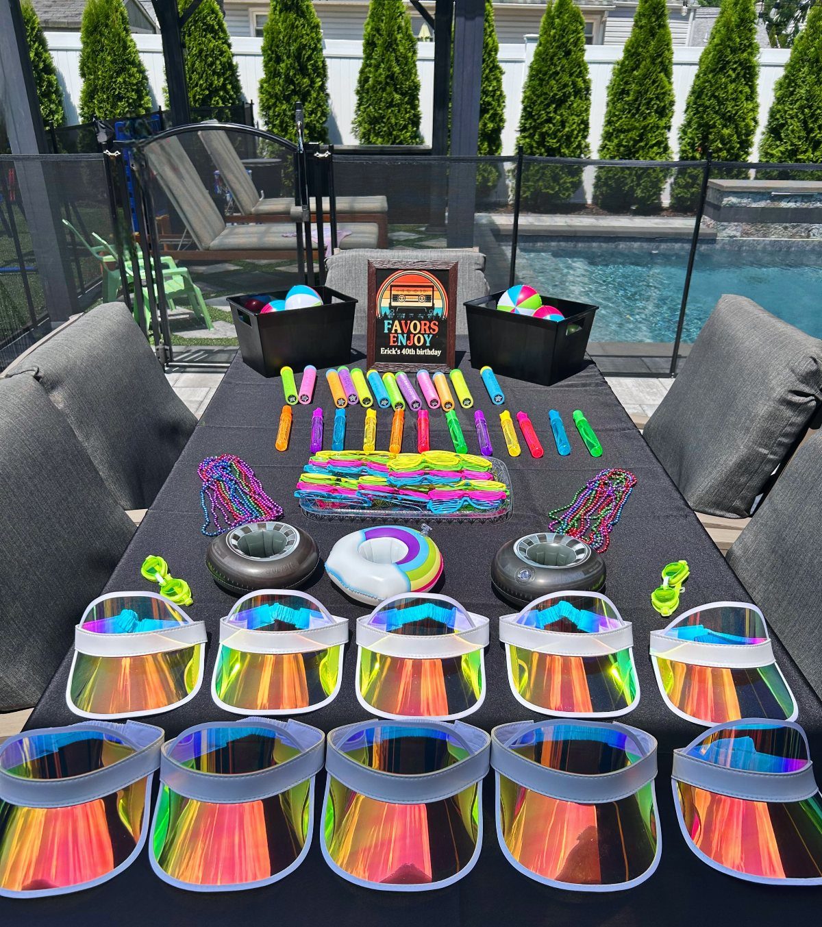 Rainbow-colored party favors and iridescent visors on a table by a swimming pool.