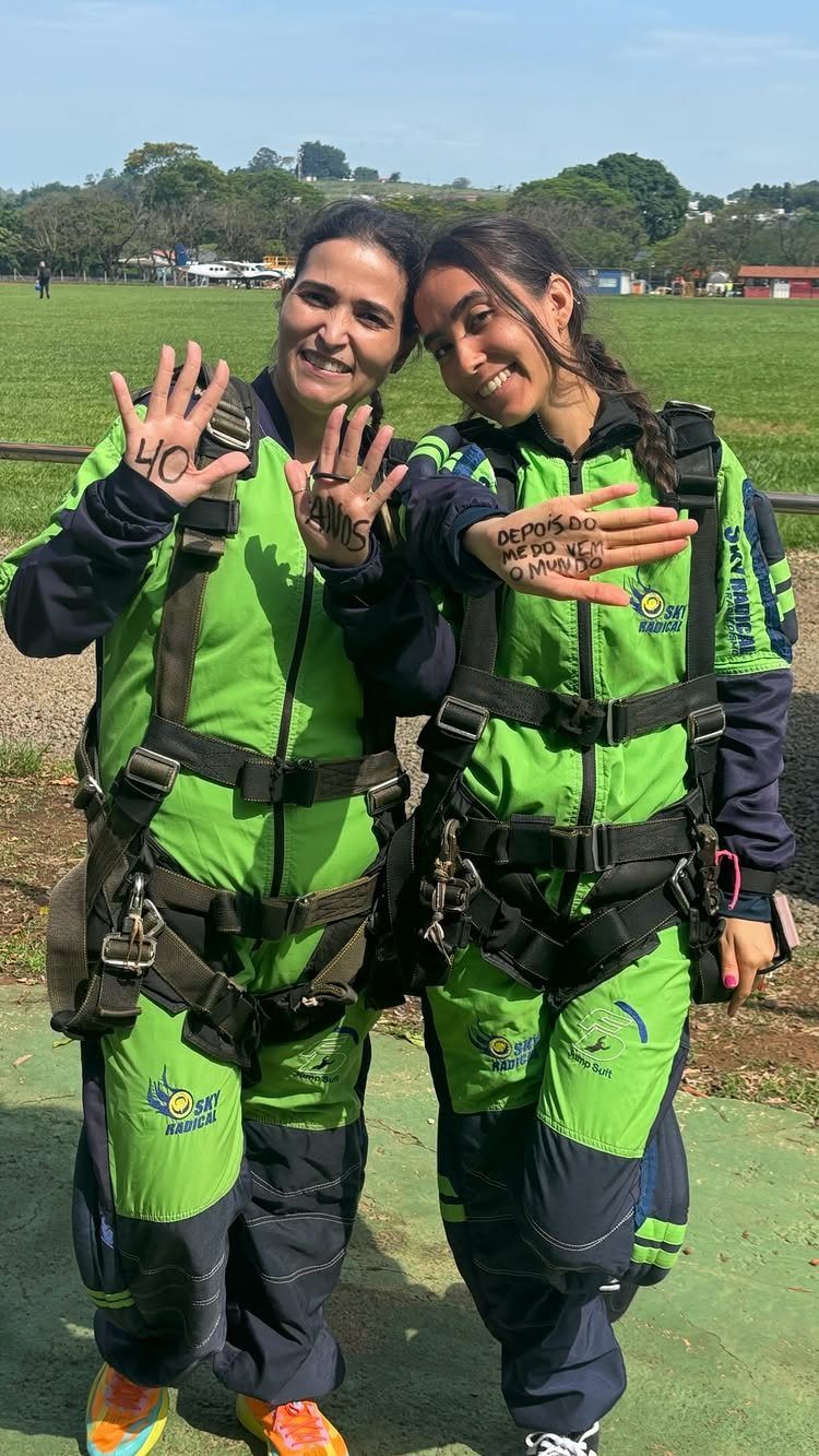 Two women in skydiving gear posing outdoors, one showing 40 on her palm.