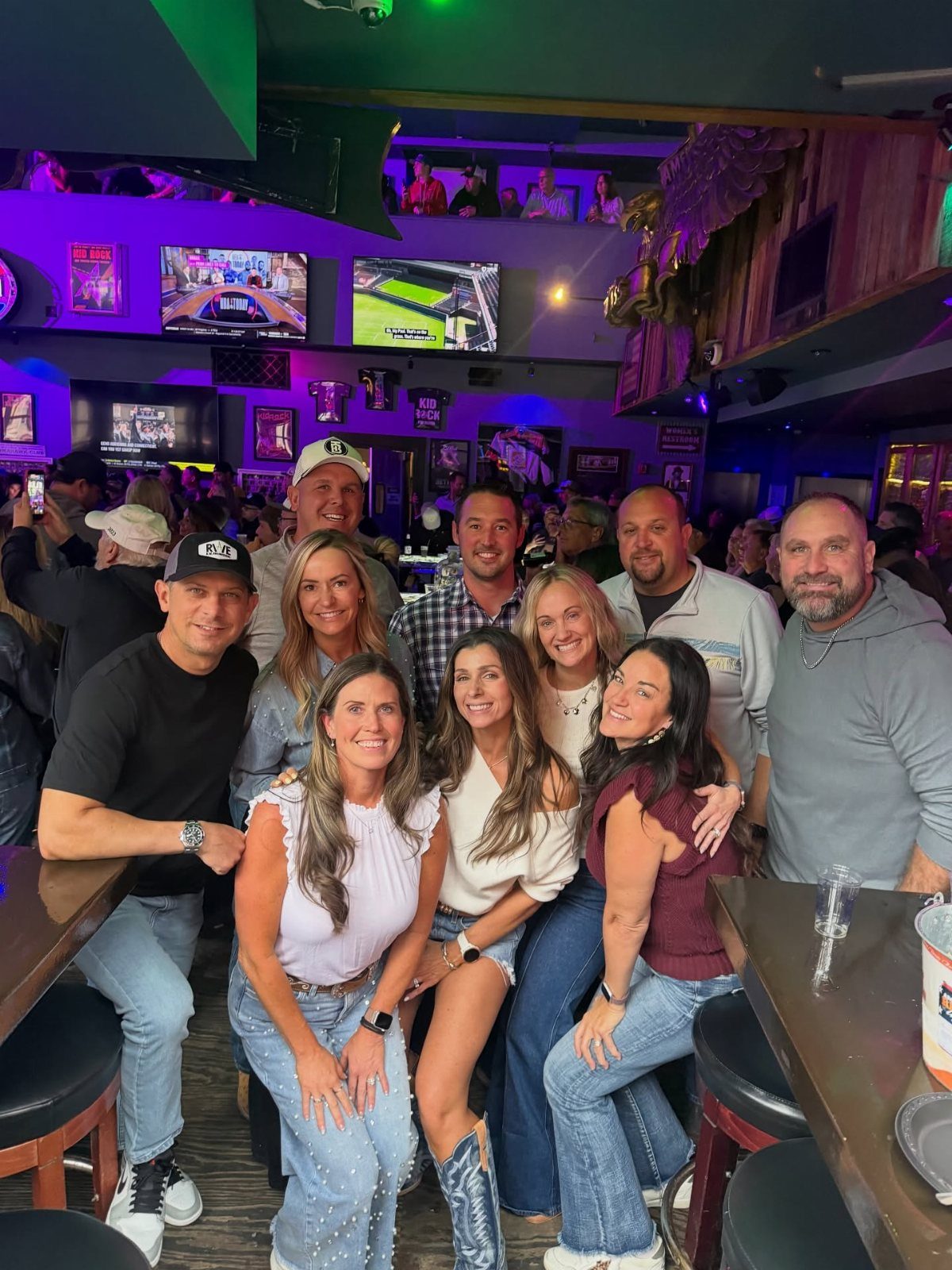 Group of smiling adults posing together inside a sports bar with neon lighting.