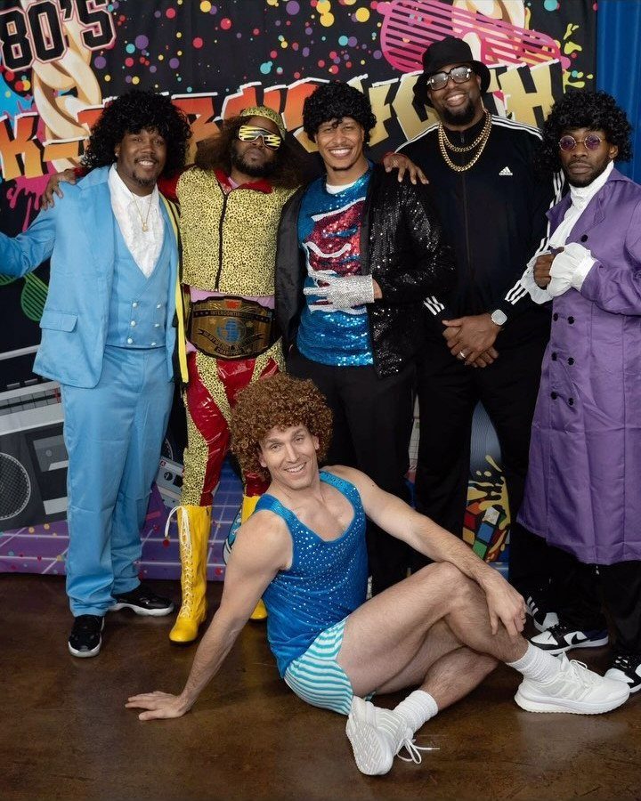 Five men in 1980s costumes posing in front of a colorful retro backdrop.