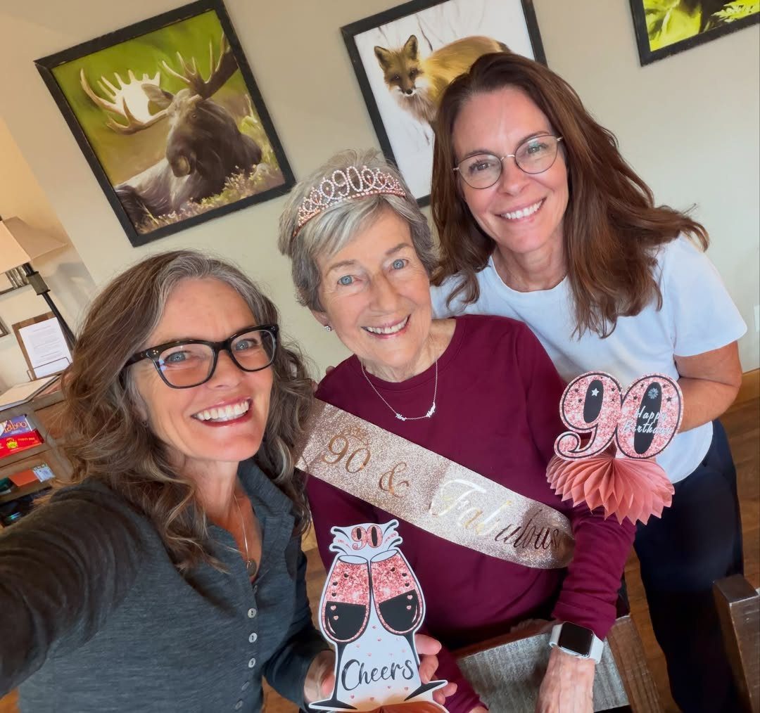 Three women smiling, with the elderly woman wearing a 90th birthday sash and tiara.