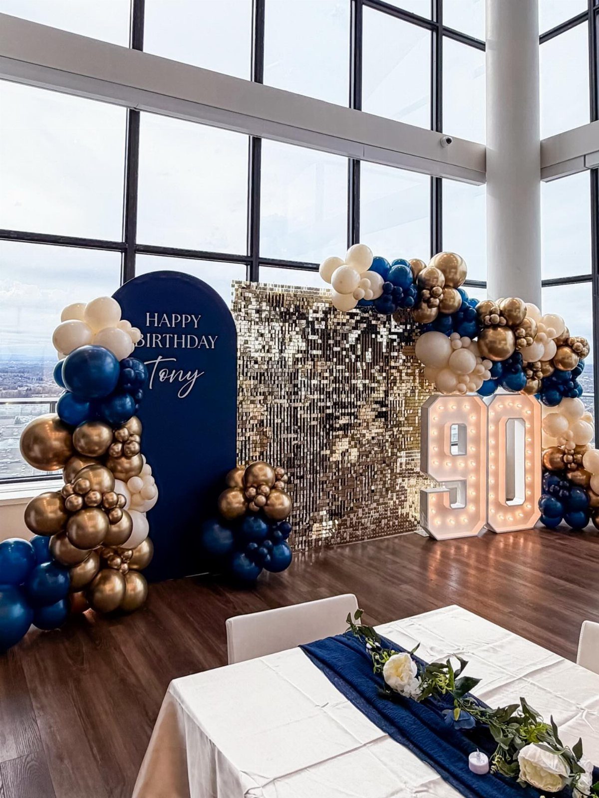 Birthday party decor with gold sequin wall, illuminated 90 numbers, and navy balloons.