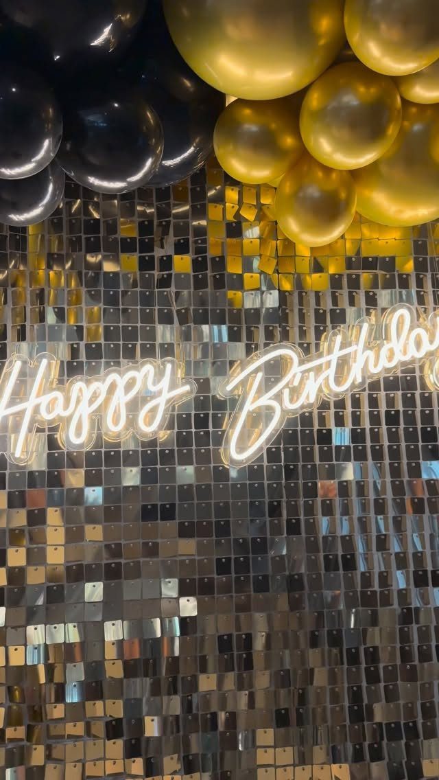 Gold and black balloons above a silver sequin wall with Happy Birthday neon sign