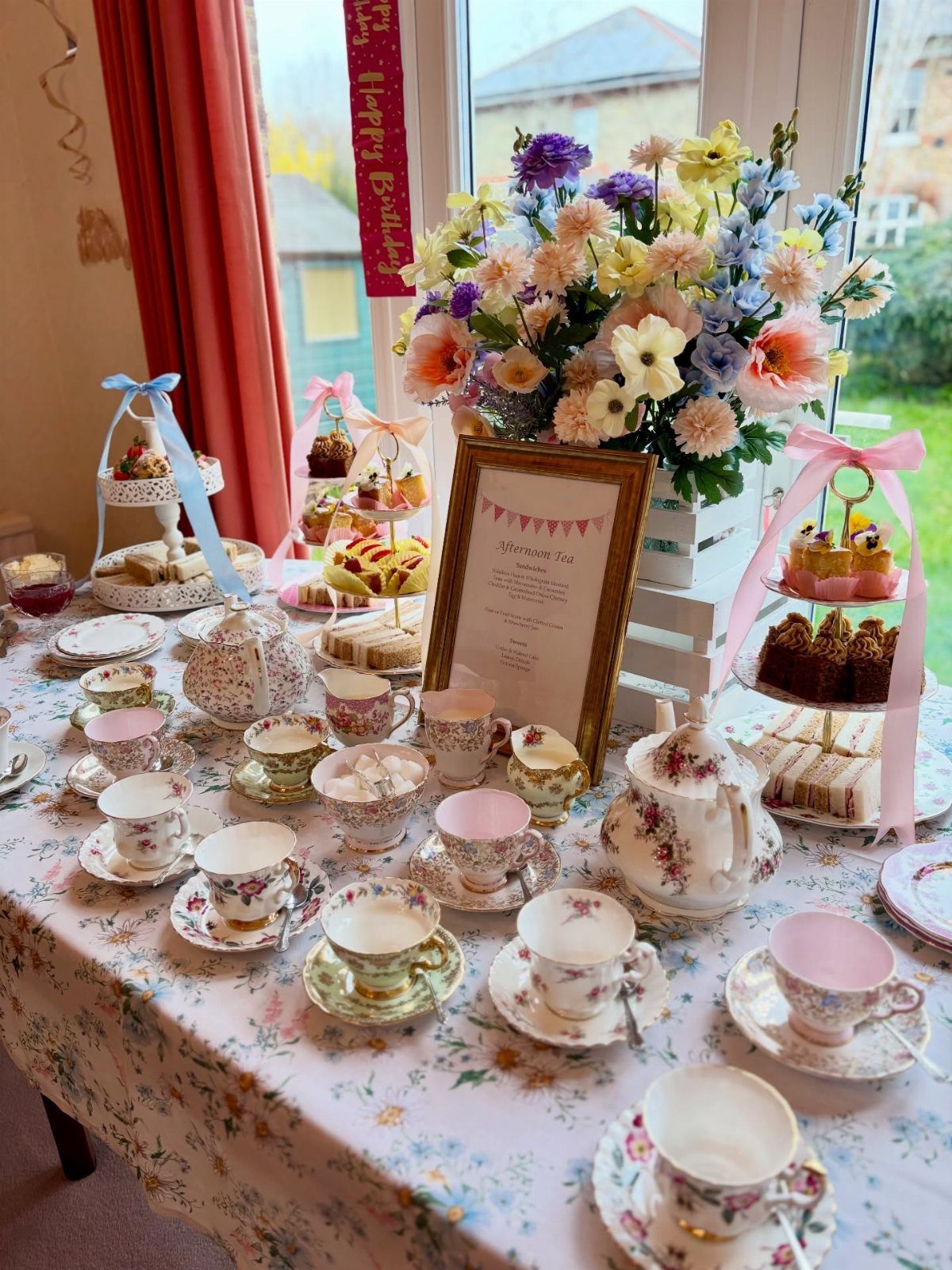 Vintage tea party table setting with floral teacups, tiered cake stands, and colorful flowers.