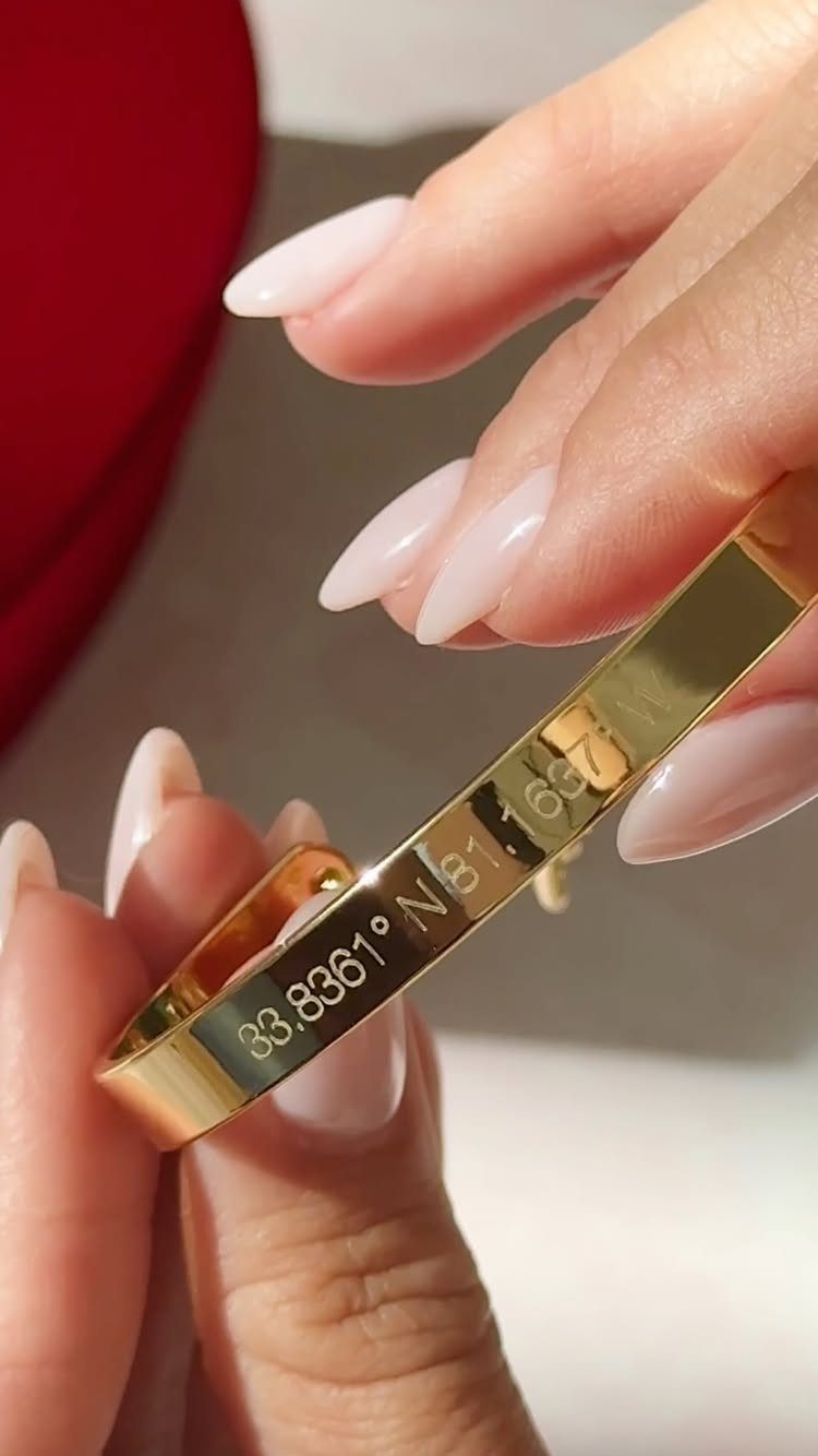Gold ring with engraved coordinates held by hands with pink manicured nails