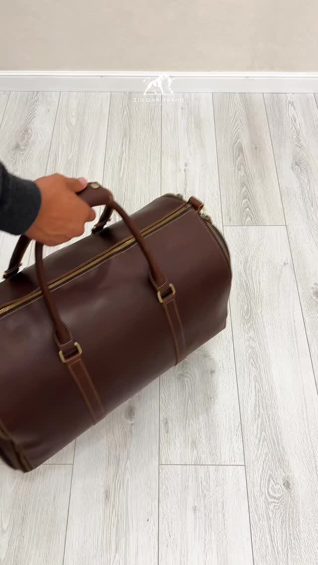 Brown leather duffel bag held by a hand on a light wood floor.