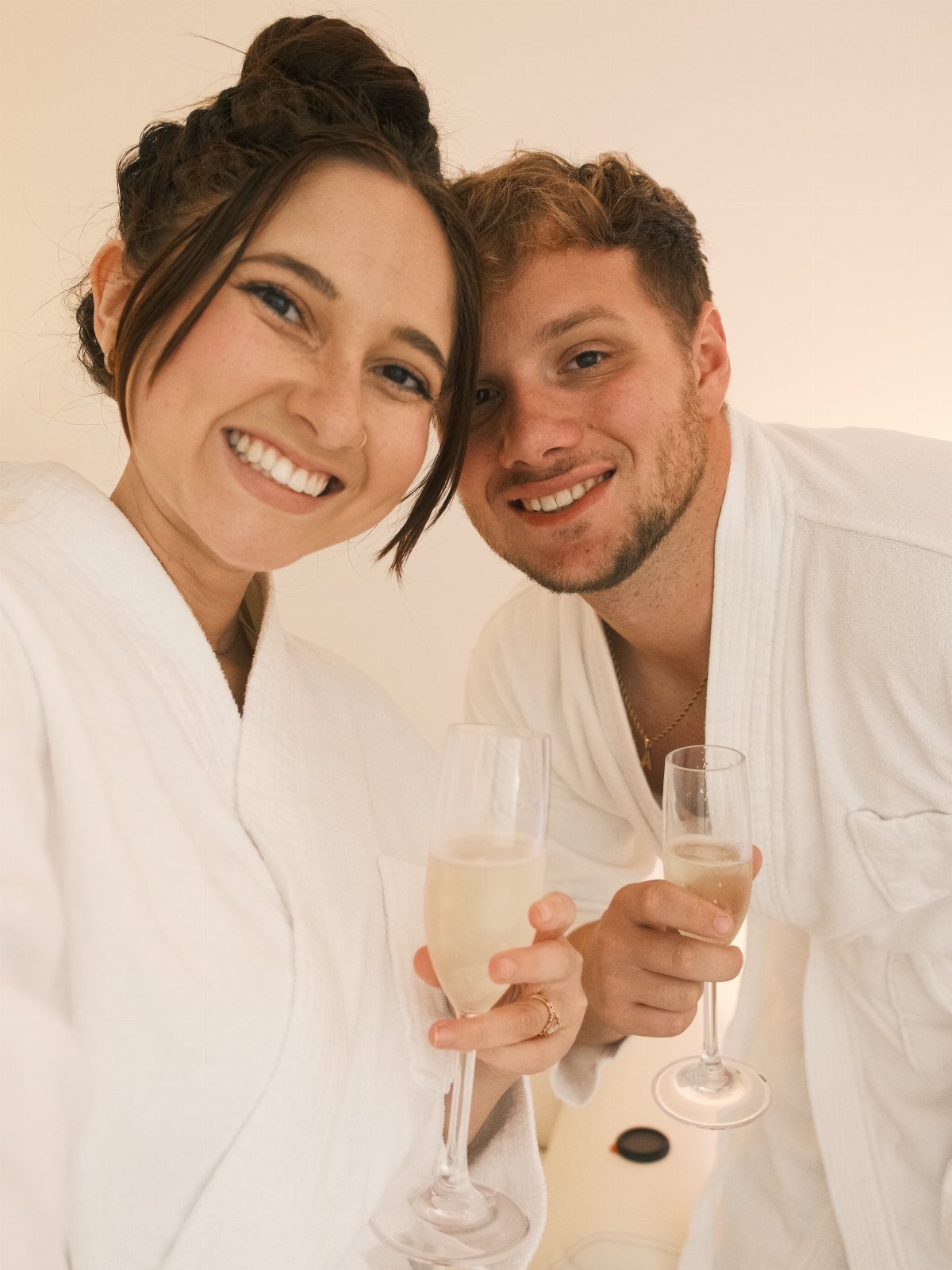 Couple in white robes smiling and holding champagne glasses during a spa day.