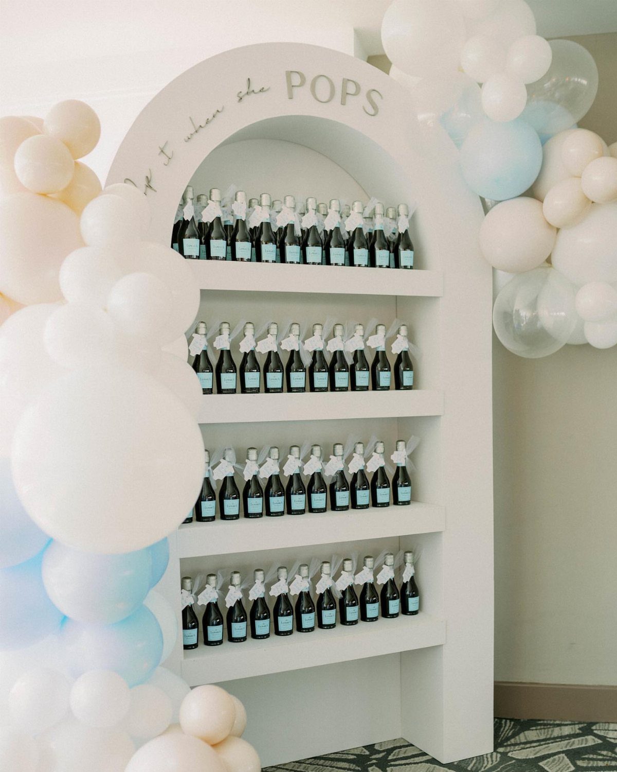 Mini bottles of wine with white ribbons on a white arched shelf with balloons