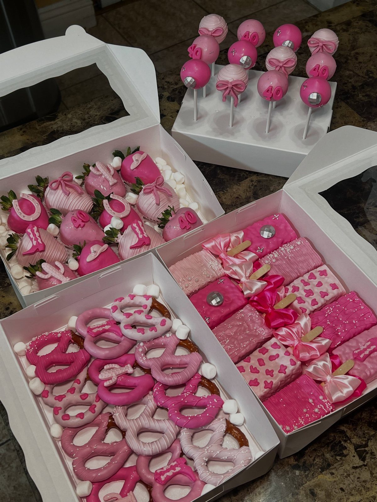 Assorted pink chocolate-covered strawberries, pretzels, and cake pops in white gift boxes.