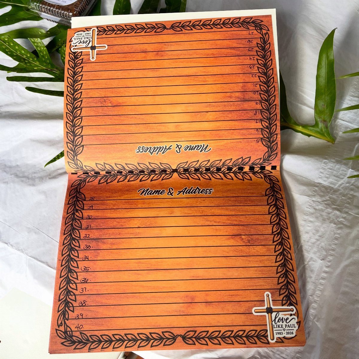 Open memorial guest book with orange pages, leaf borders, and a cross emblem.