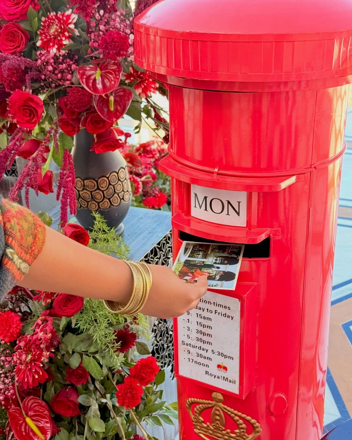 Hand inserting a photo card into a red Royal Mail pillar box among flowers