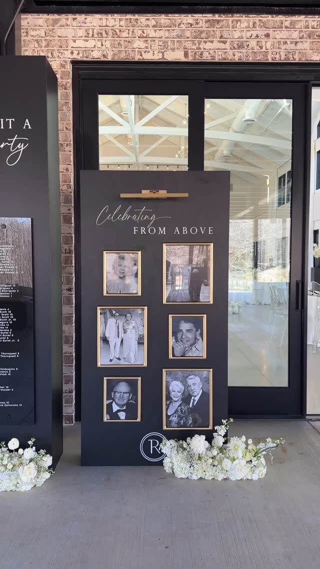 Black memorial board with framed photos and gold text surrounded by white flowers.