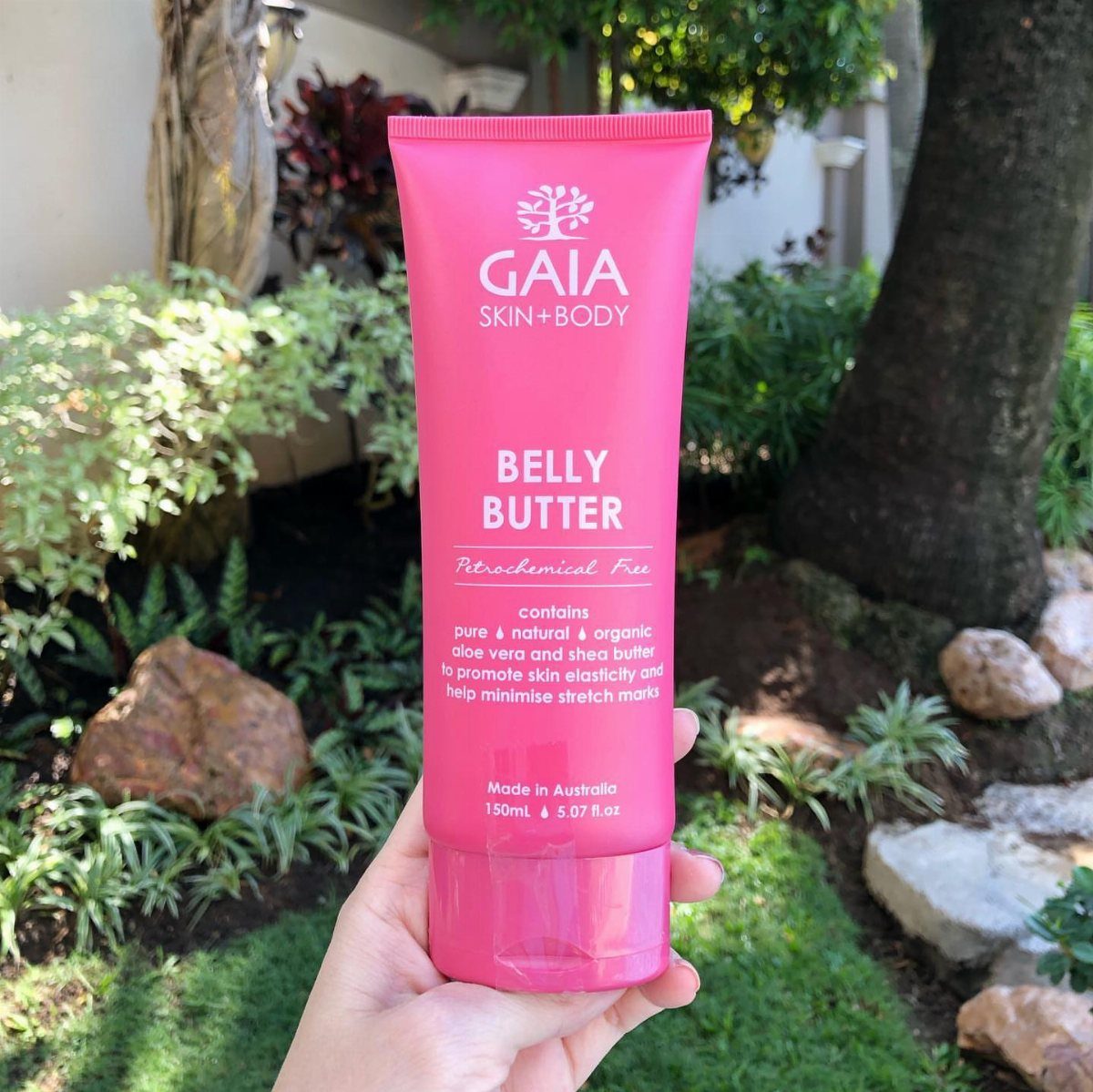 Pink tube of Gaia Belly Butter held in a hand outdoors among greenery.