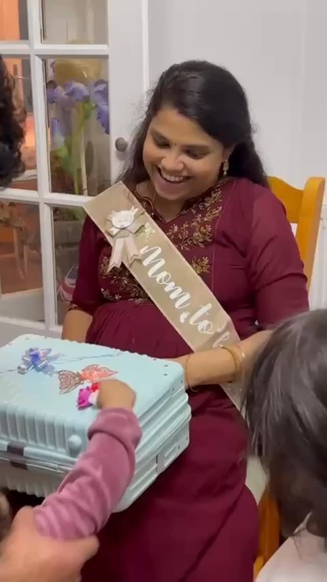 Pregnant woman in maroon dress wearing a Mom to be sash smiling indoors