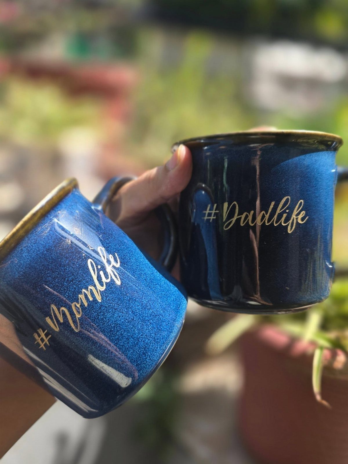 Two blue glittery mugs with gold #Momlife and #Dadlife text held outdoors.