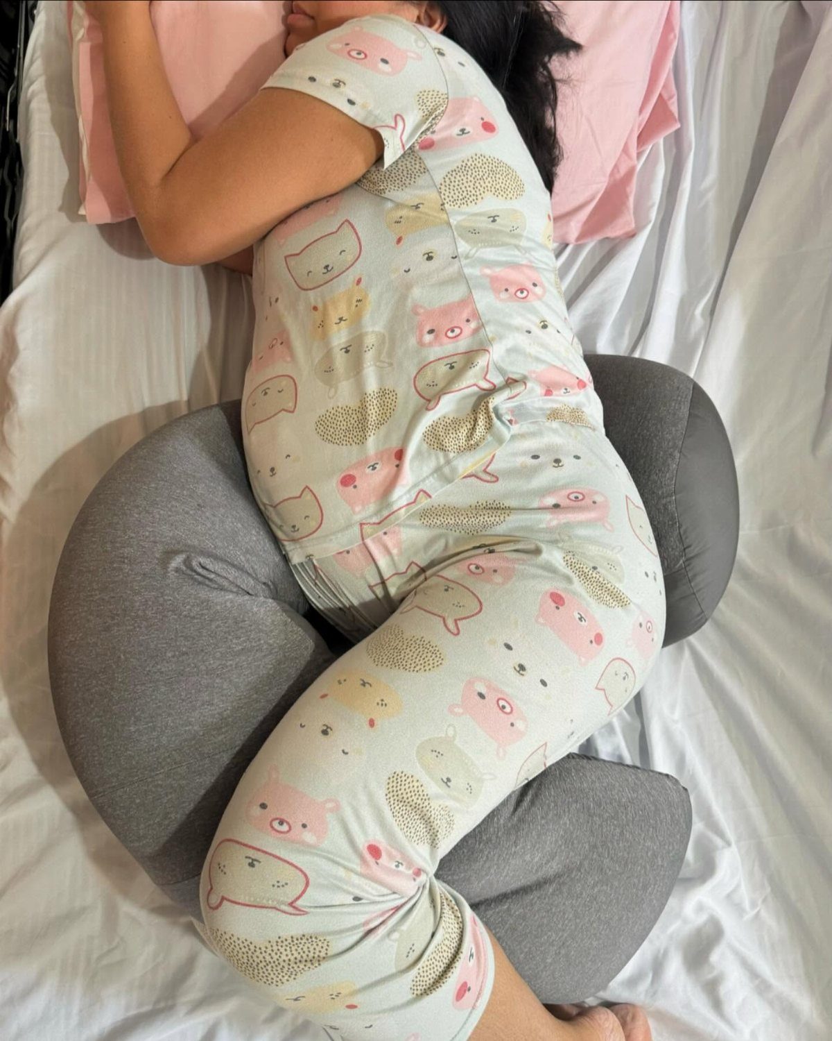 Pregnant woman in pajamas resting on a grey U-shaped pregnancy pillow on a bed
