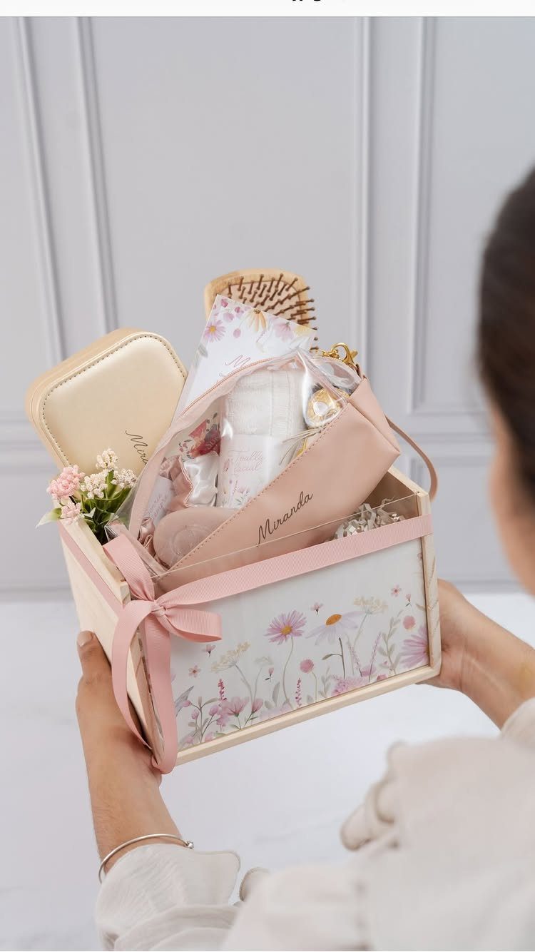 Floral gift box filled with beauty products and a pink ribbon held by hands.