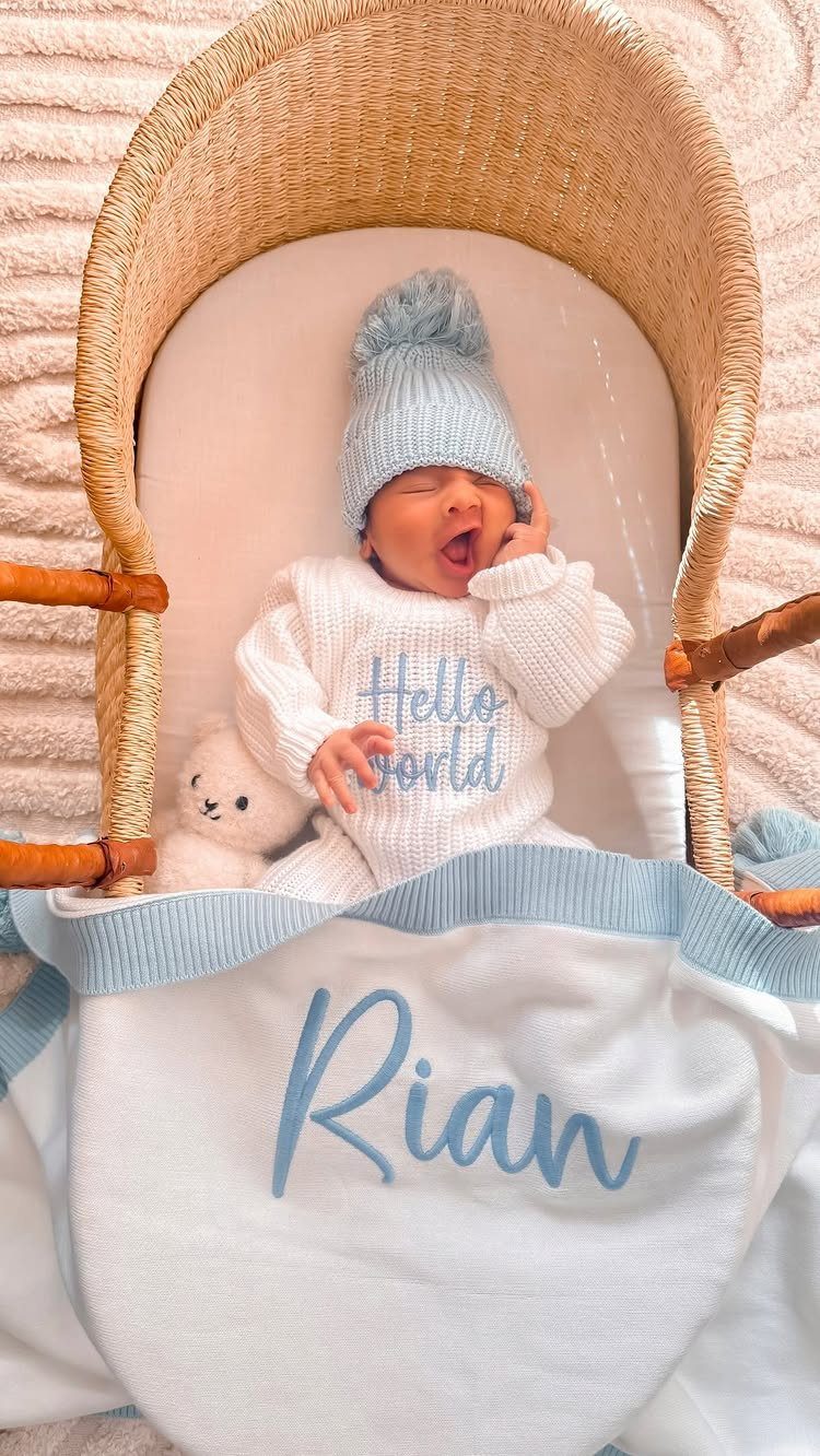 Newborn baby in a white Hello World sweater and blue beanie in a bassinet