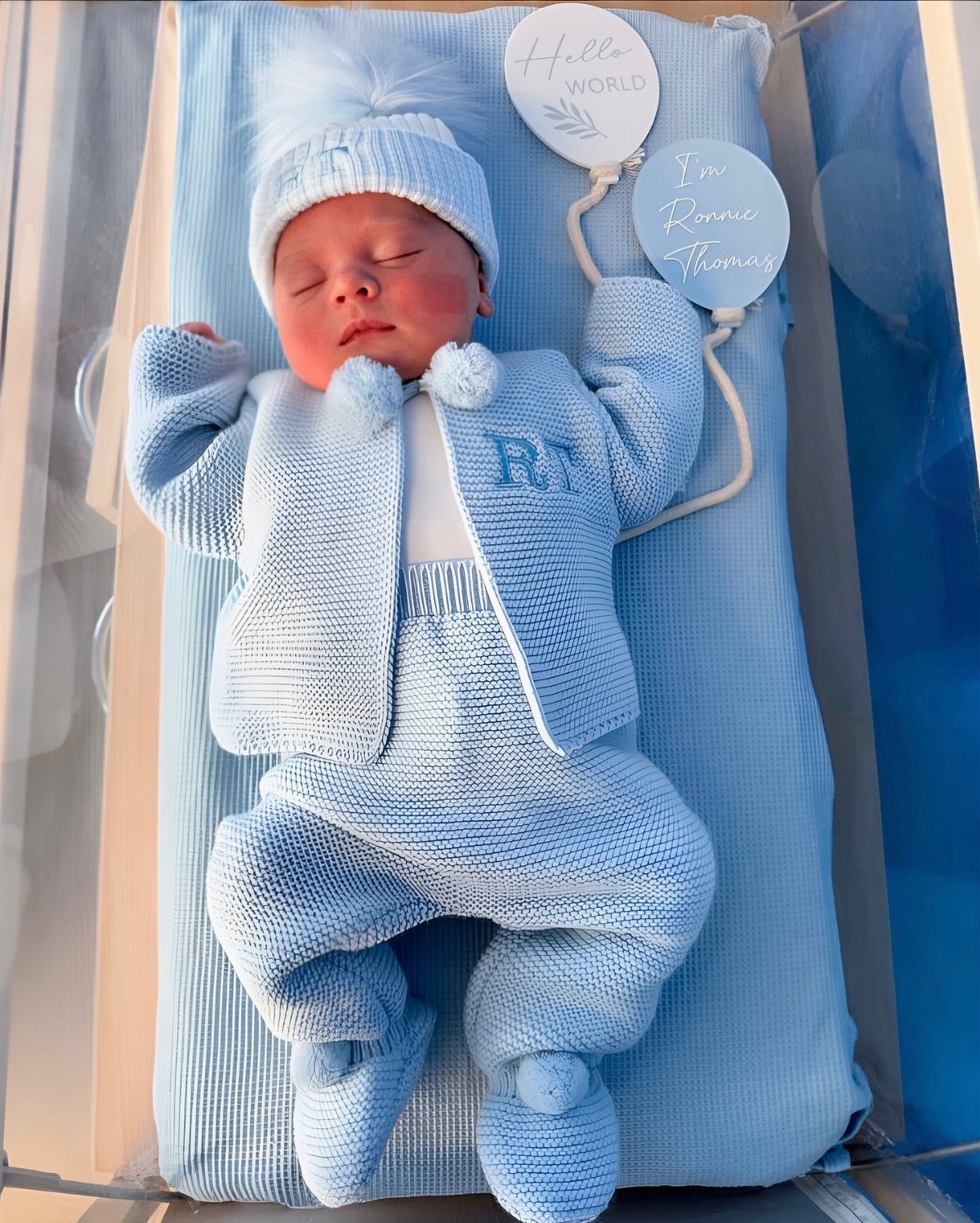 Sleeping newborn baby in a light blue knitted outfit inside a hospital bassinet.