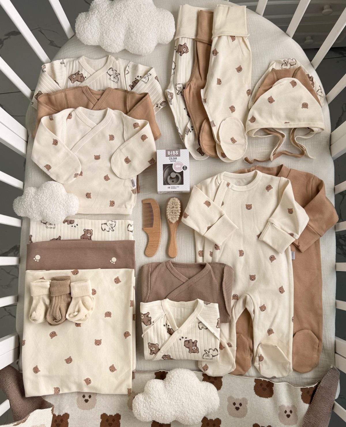 Newborn baby clothes in cream and tan tones arranged in a white crib.