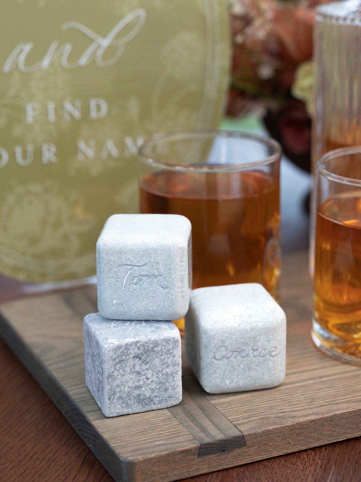 Engraved grey whiskey stones stacked on a wooden board next to amber drinks.