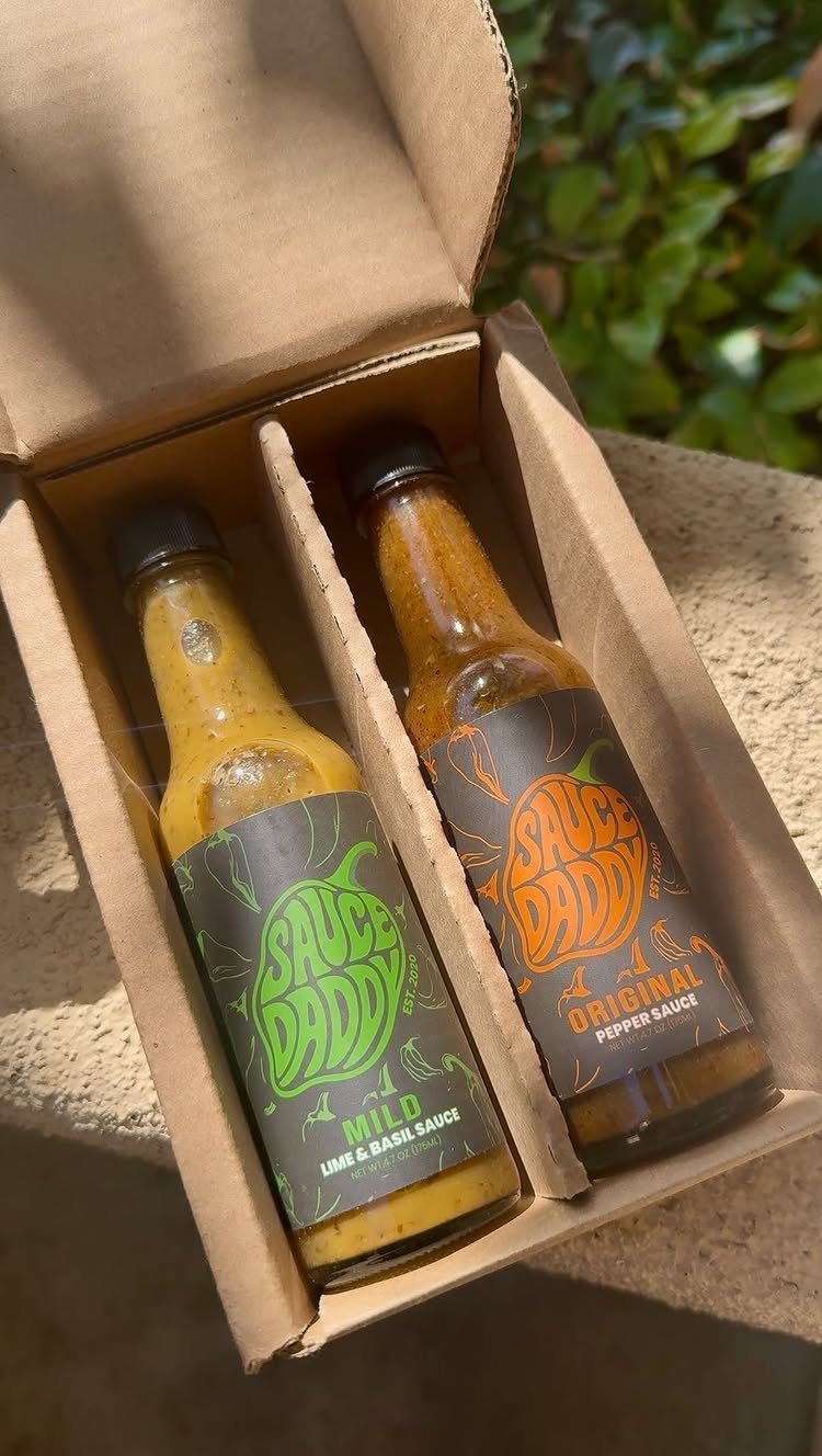 Two bottles of hot sauce in a brown cardboard gift box outdoors.
