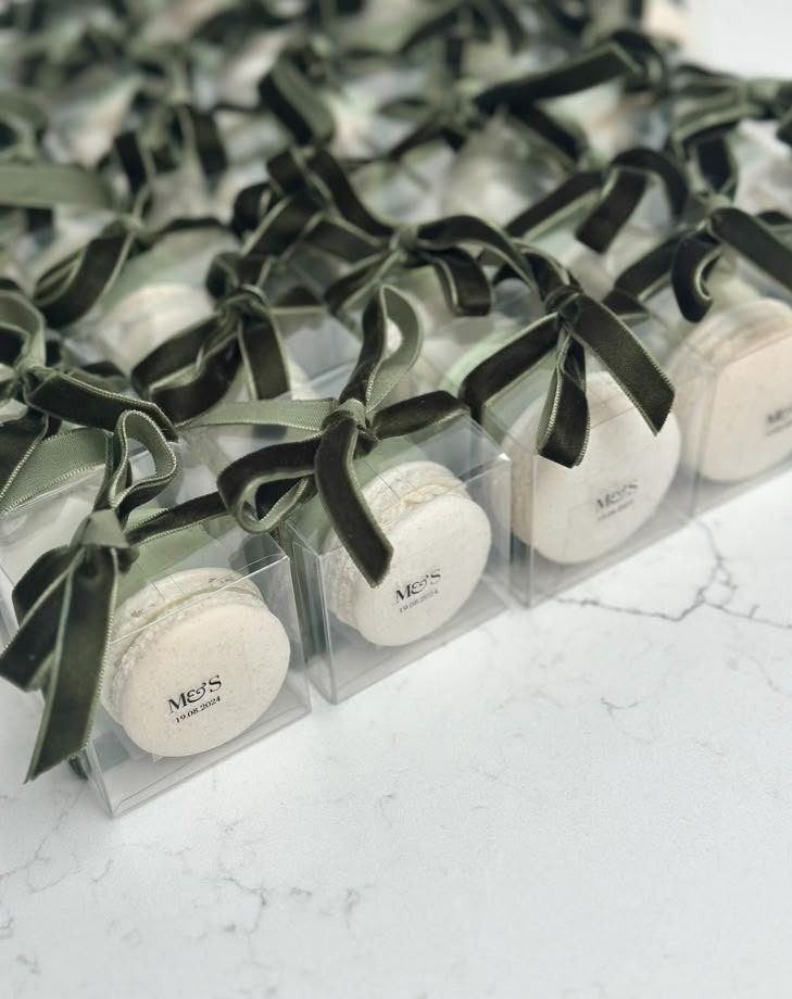 White macarons in clear boxes tied with olive green velvet ribbons on marble.