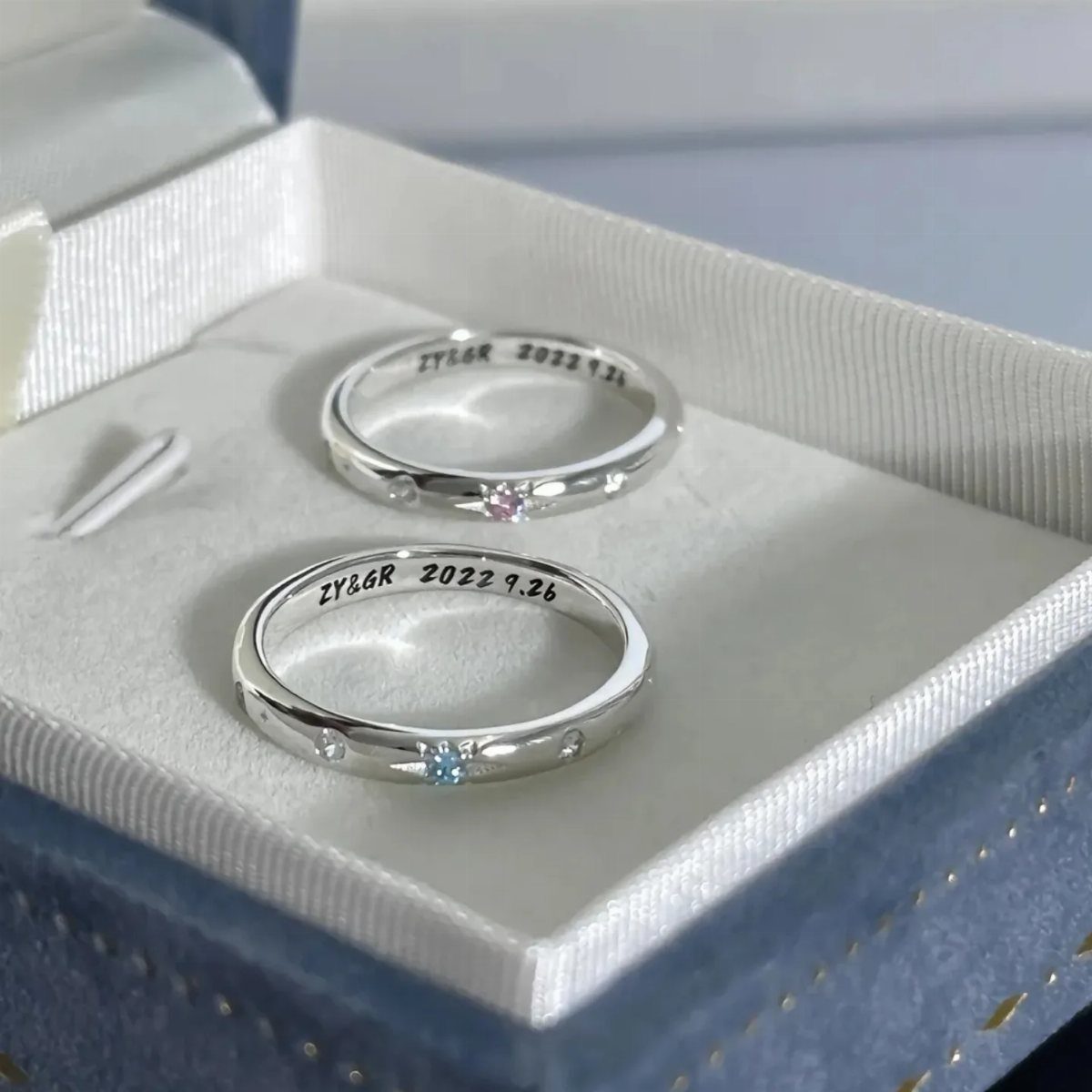 Two engraved silver rings with small gemstones inside a blue velvet jewelry box.