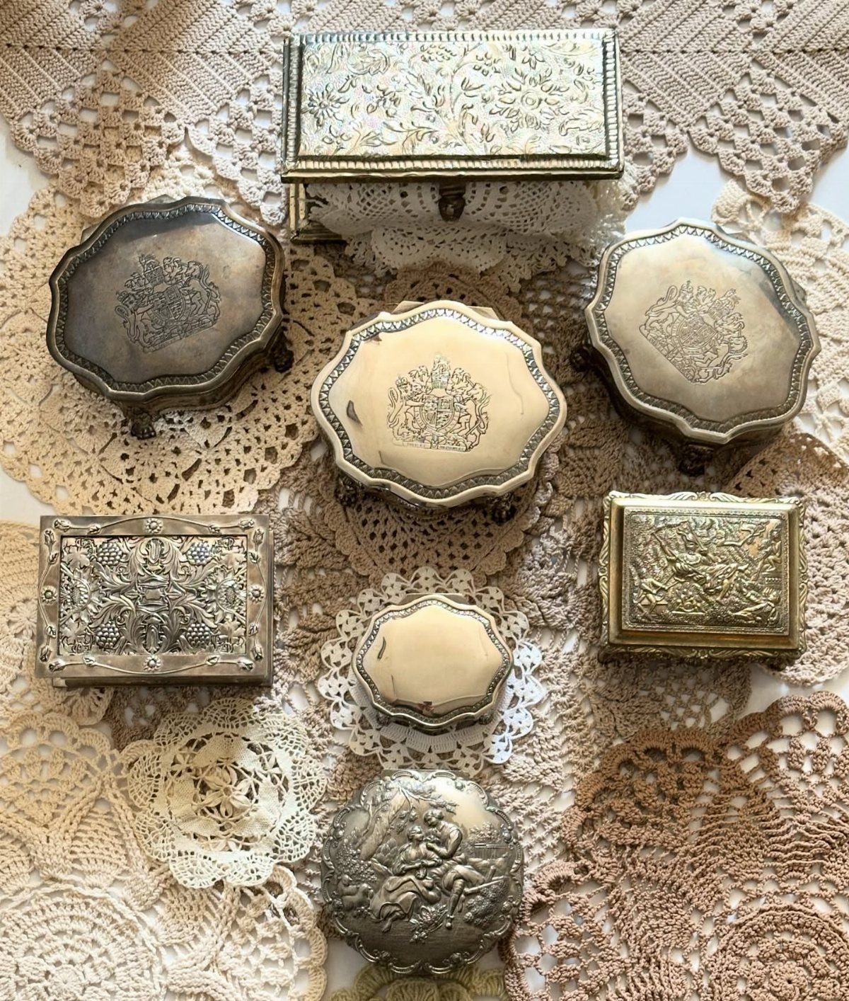 Various ornate silver jewelry boxes arranged on delicate cream and beige lace doilies.