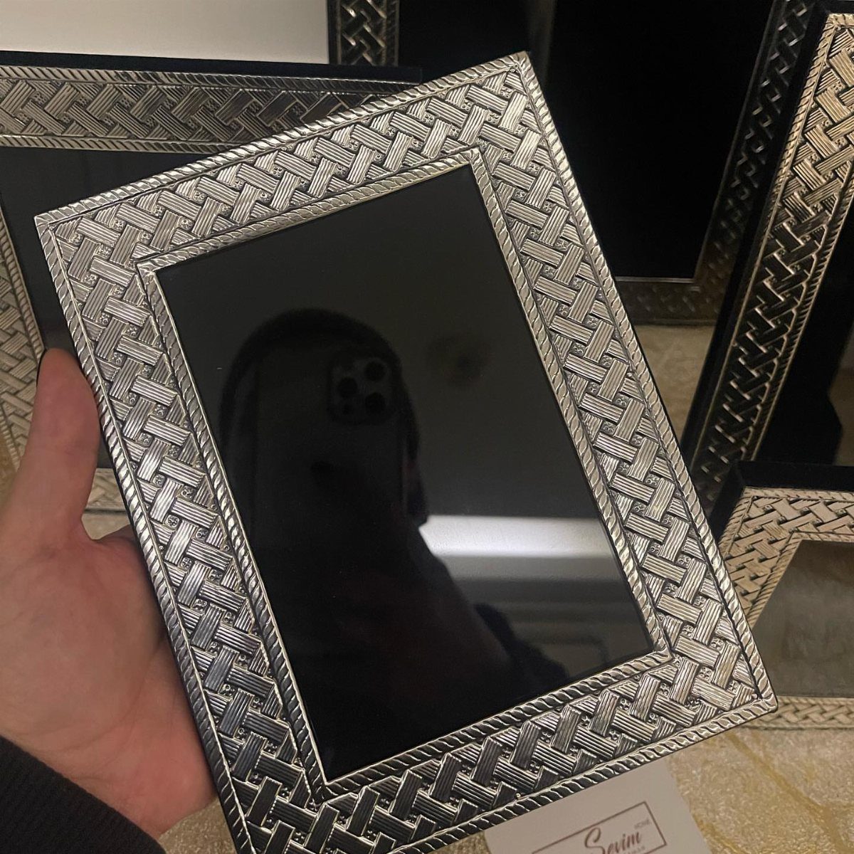 Silver woven picture frame held by a hand, reflecting a person with a phone.