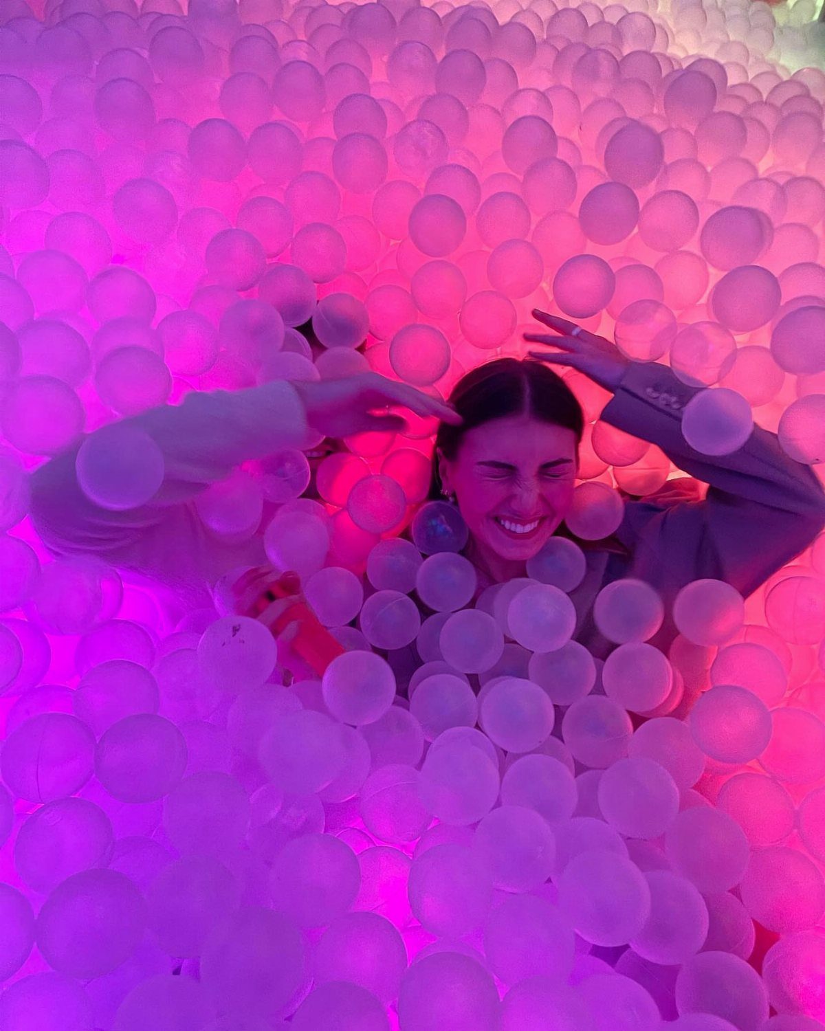 Young woman laughing while playing in a pink illuminated ball pit.