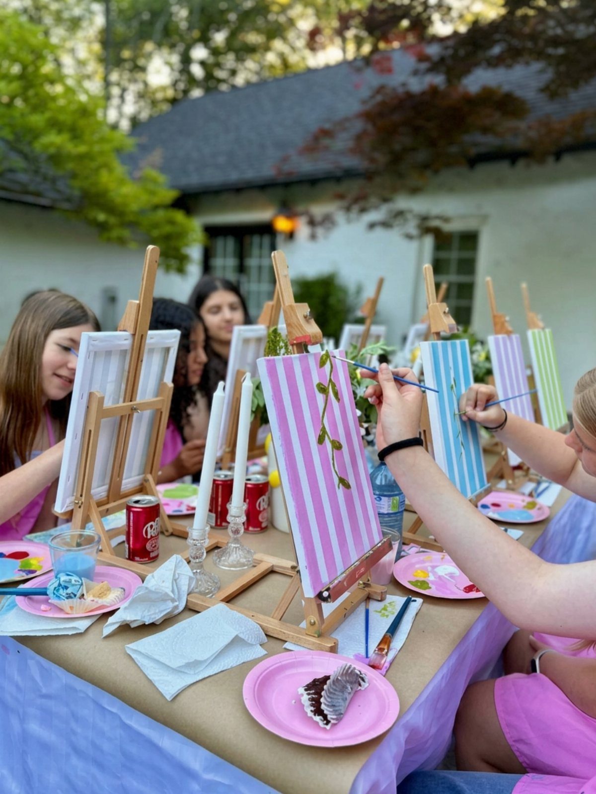 Teenage girls painting colorful striped canvases on wooden easels at an outdoor party.
