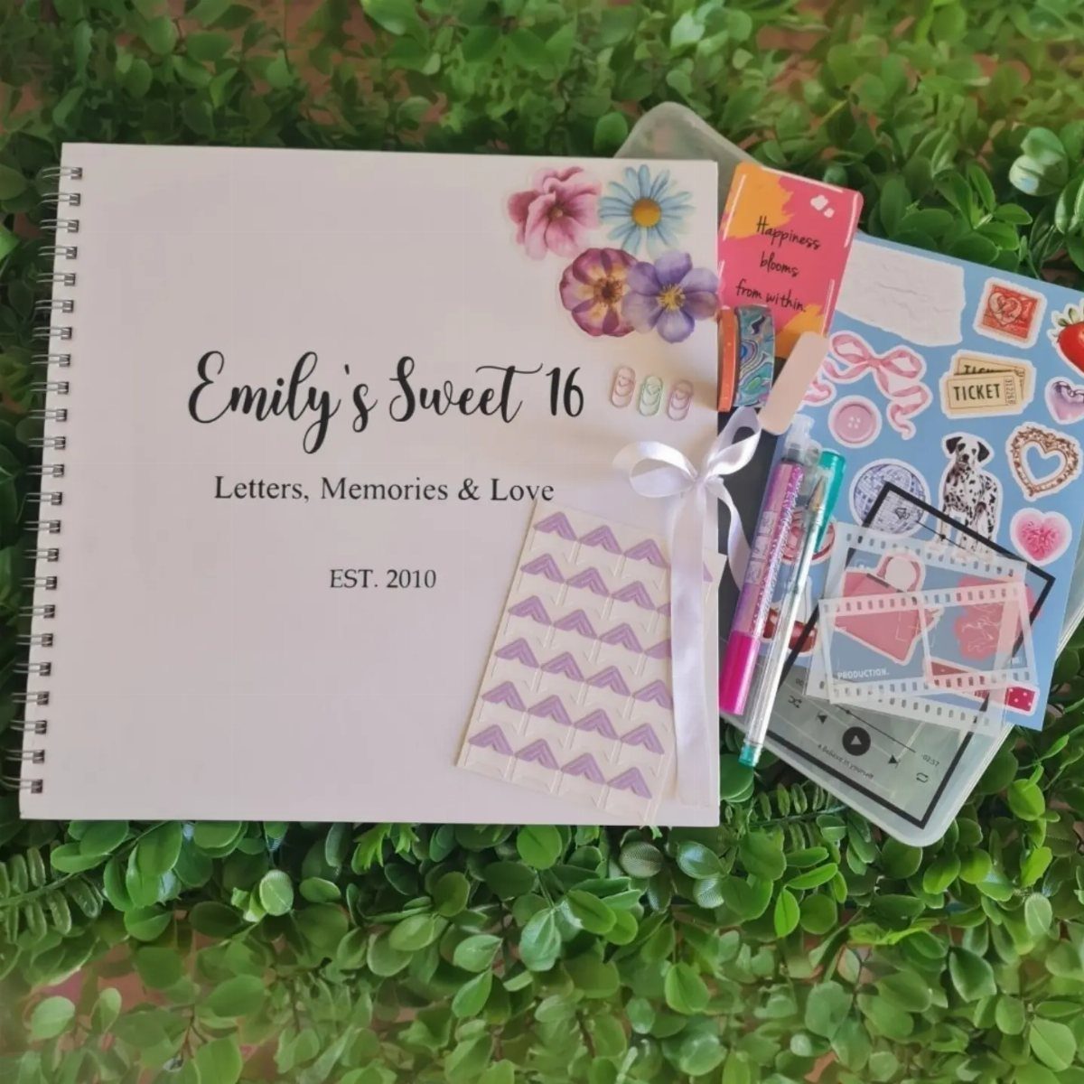 Personalized Sweet 16 notebook with floral cover and stationery on green foliage.