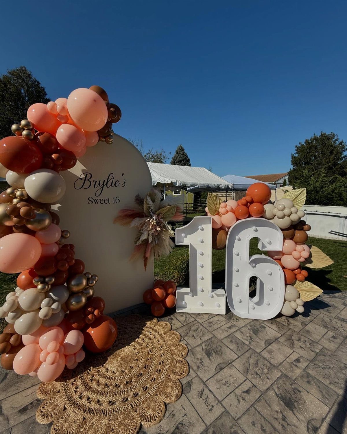 Sweet 16 party backdrop with peach balloons, illuminated numbers, and boho jute rug.
