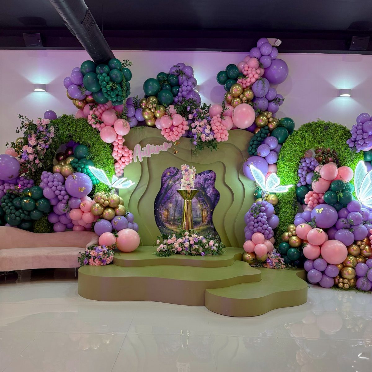 Party backdrop with purple and pink balloons, greenery, and glowing fairy wings.
