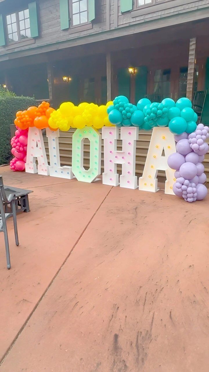 Large ALOHA marquee letters with colorful balloon arches on a concrete patio.