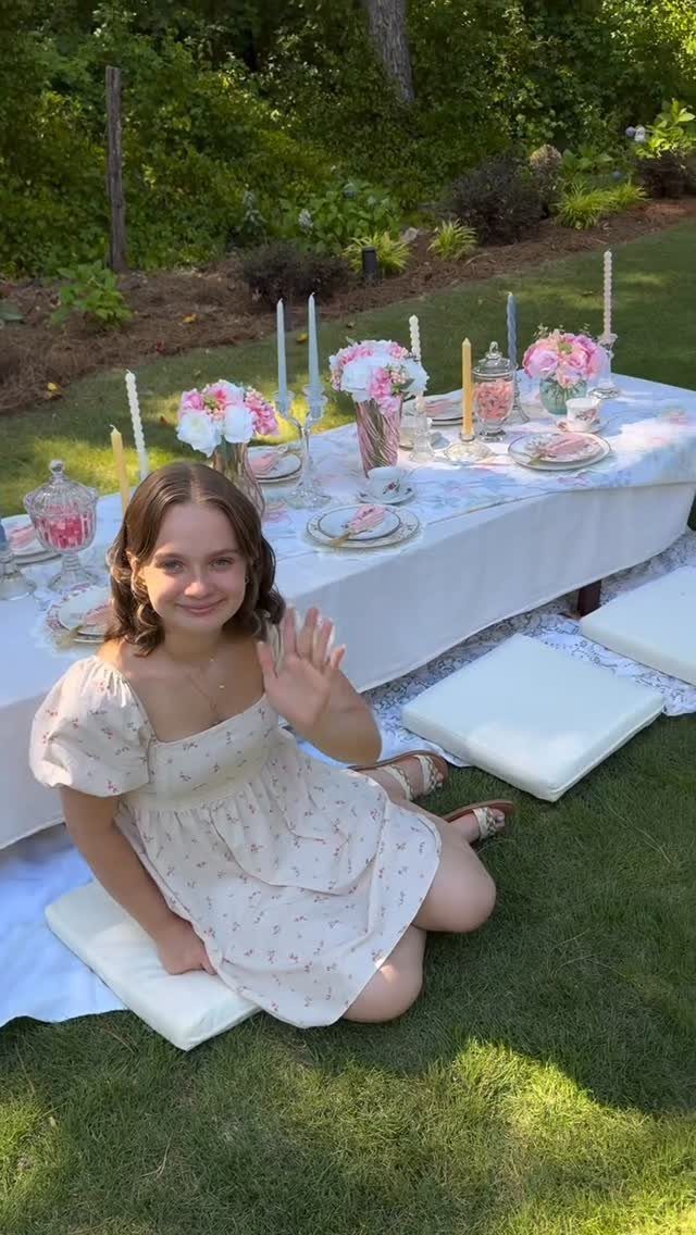 Young woman in floral dress sitting by a decorated garden tea party table.